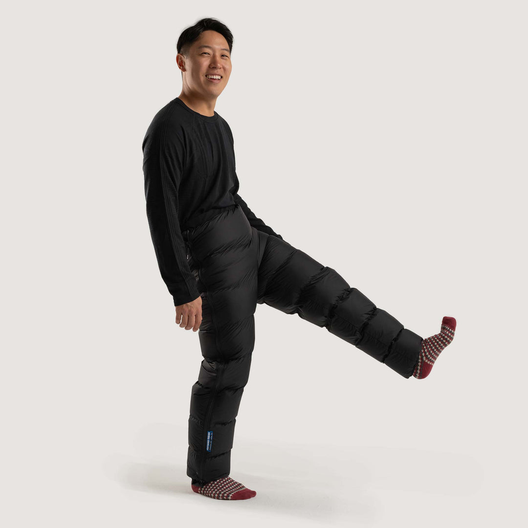 Studio front view of a man wearing a black Feathered Friends Helios down pants with one leg up