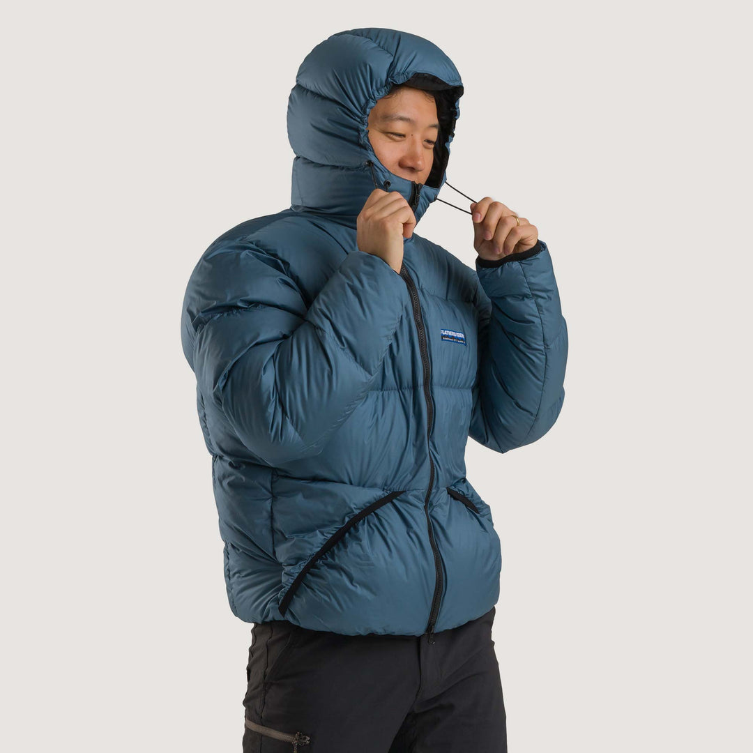 Studio front hood-detail view of a man wearing a slate blue Feathered Friends Hooded Helios down puffer jacket