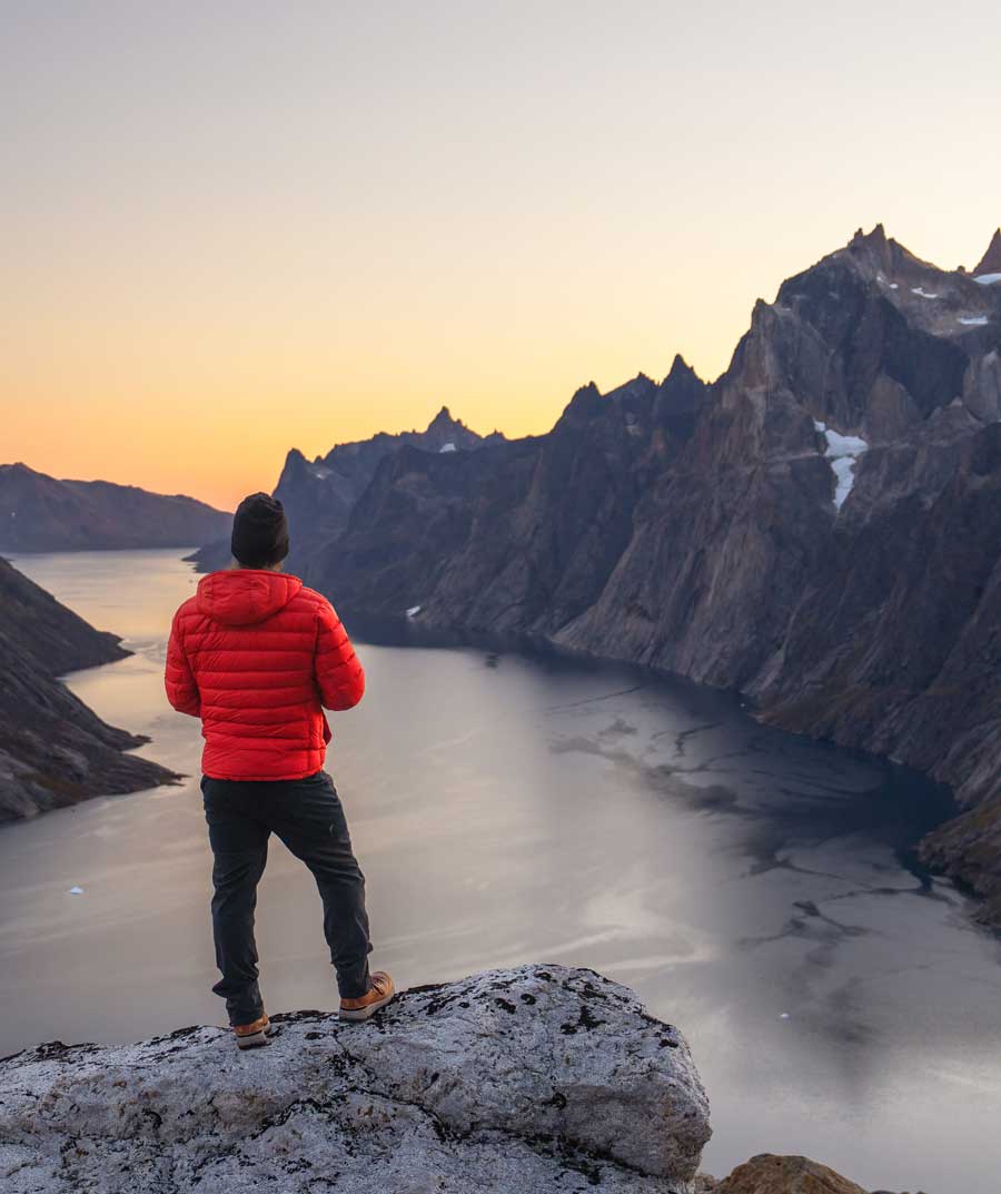 Person in a red Feathered Friends Eos jacket standing on a mountain overlooking a fjord at sunset