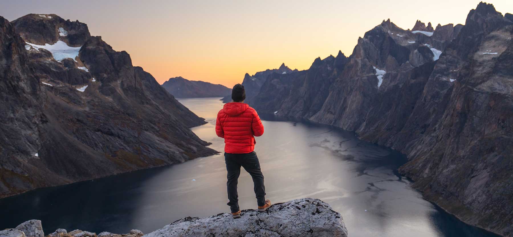 Person in a red Feathered Friends Eos jacket standing on a mountain overlooking a fjord at sunset